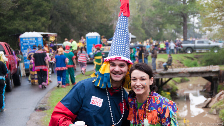 Smiling couple in colorful Mardi Gras clothing pose beside a wet road and festival crowd.