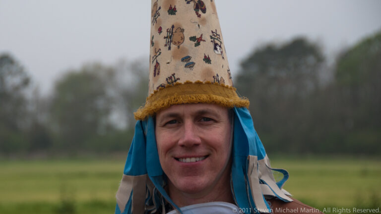 Smiling man in a tall patterned hat with blue ribbons stands before a blurred green field.