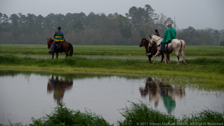 Two costumed riders on horses cross shallow water, with reflections visible below them.