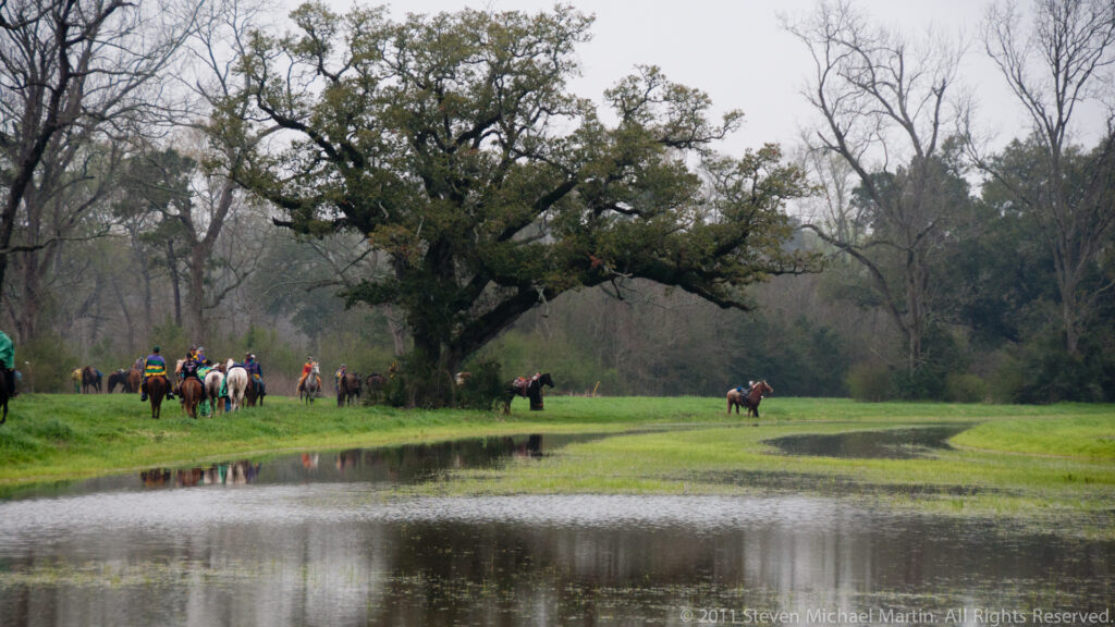 Costumed riders pass beneath a broad oak beside reflective water and green pasture.