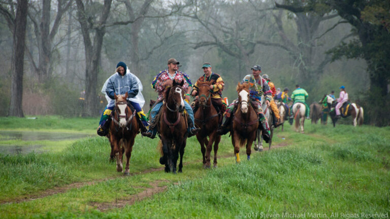 Costumed horsemen ride toward the camera along a muddy track, with more riders behind them.