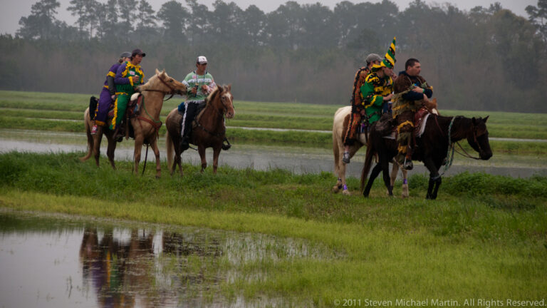 Costumed riders pause on horses beside shallow water, with one person holding a small flag.