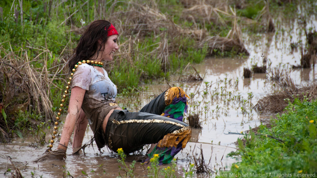 Woman wearing beads and a red head covering sits in a muddy, plant-lined ditch.