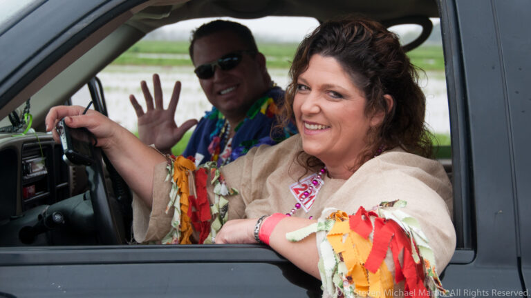 Smiling woman with beads leans from a vehicle window while a man behind her waves.