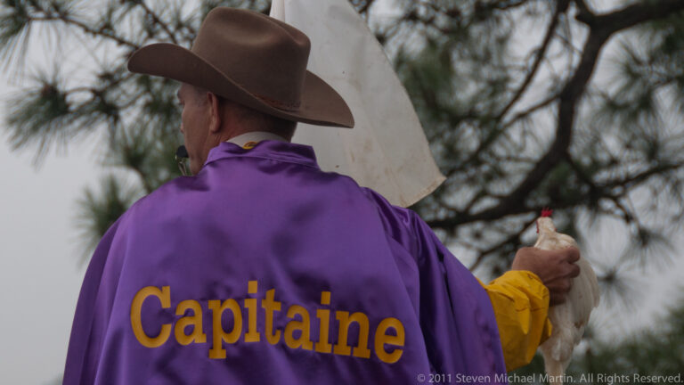 Participant in a purple “Capitaine” jacket holds a white chicken beneath bare tree branches.