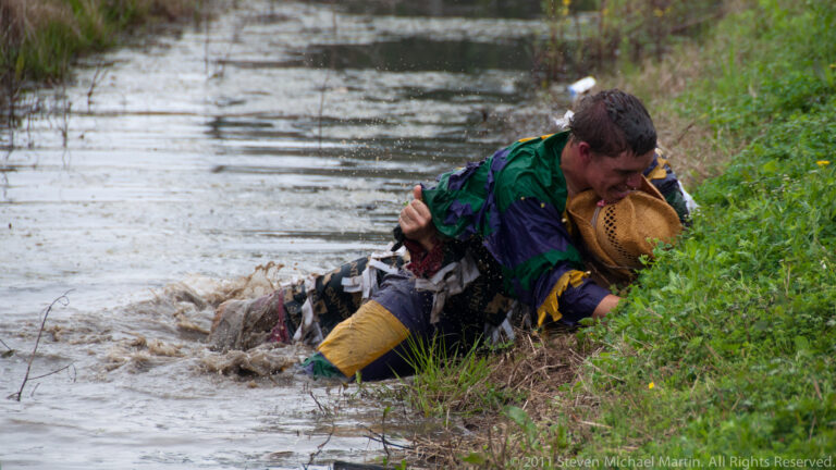 Costumed man kneels in shallow water beside another person at a grassy muddy bank.