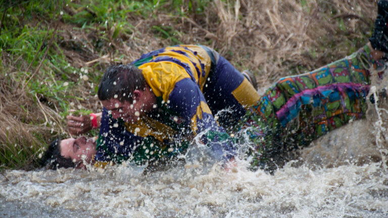 Costumed participant splashes into shallow water over another person in a reed-lined ditch.