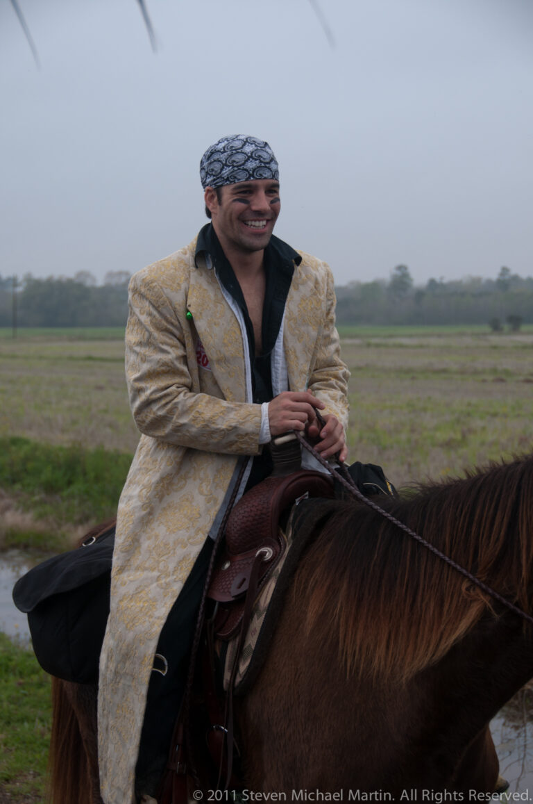 Smiling man in an embroidered light coat and patterned head wrap sits on a brown horse.