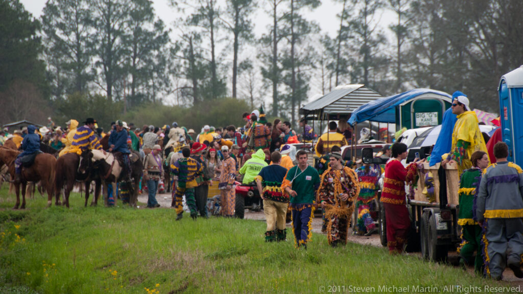 Costumed Mardi Gras participants and horseback riders gather beside parked trucks along a rural Louisiana road.