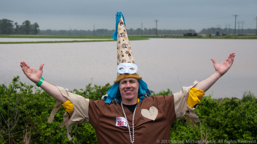 A smiling masked man in a pointed cap raises both arms beside a flooded field.