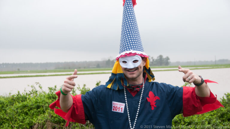 Masked man in a tall star-spangled cap and colorful tunic gestures toward the camera beside a field.
