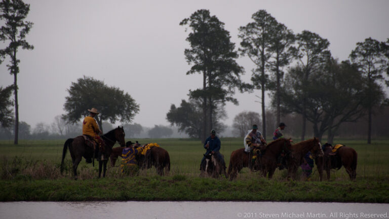 A distant line of costumed riders crosses a wet field beneath trees in dim, overcast light.