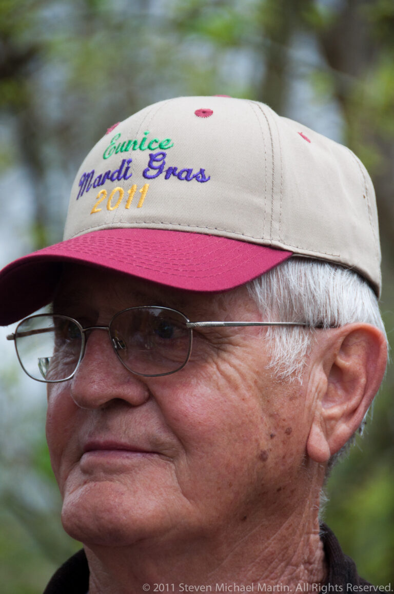 Close portrait of an older man in glasses wearing a cap embroidered Cajun Mardi Gras 2011.