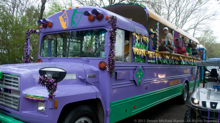 Decorated purple-and-green bus carries costumed riders along a tree-lined rural road during the parade.