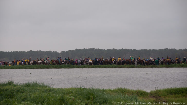 A distant line of costumed participants and horses follows the opposite bank of a wide waterway.