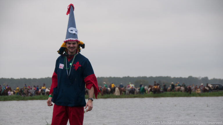 Costumed man in a pointed cap stands before a distant line of horseback riders beside water.