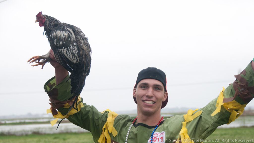 Smiling costumed participant raises a dark chicken overhead against a pale sky.