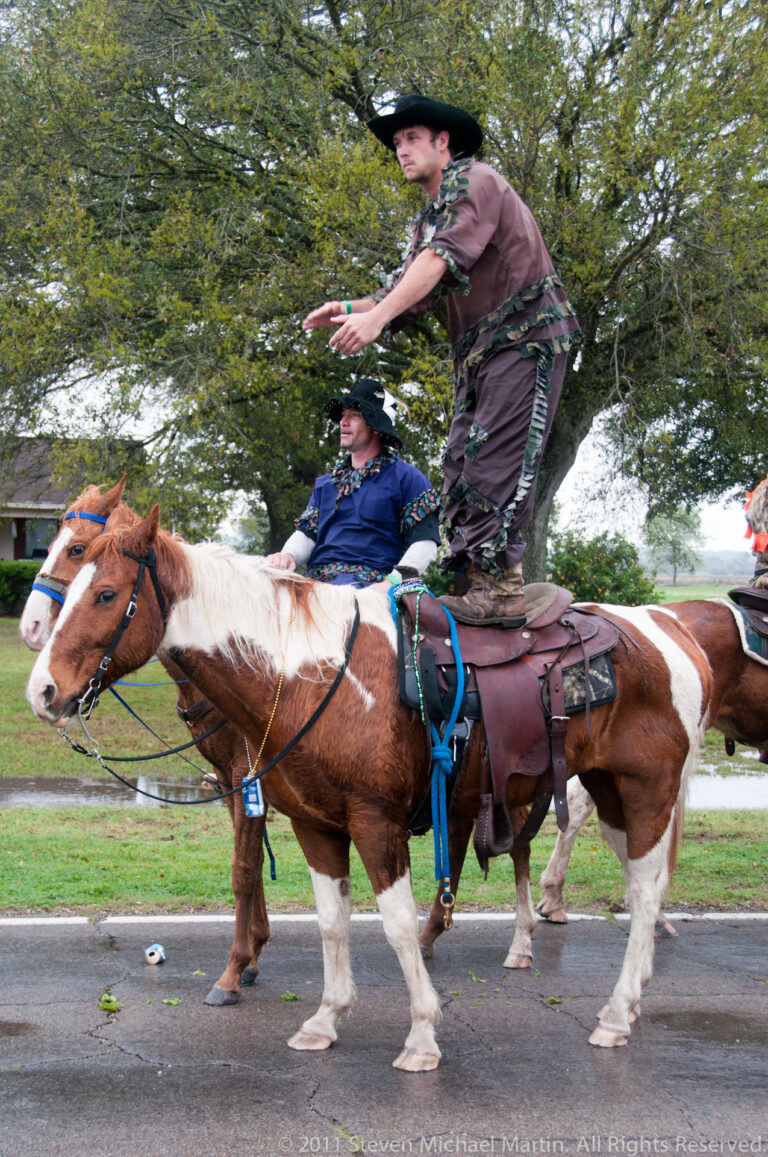 A costumed man stands on a pinto horse beside another mounted rider on a tree-lined road.
