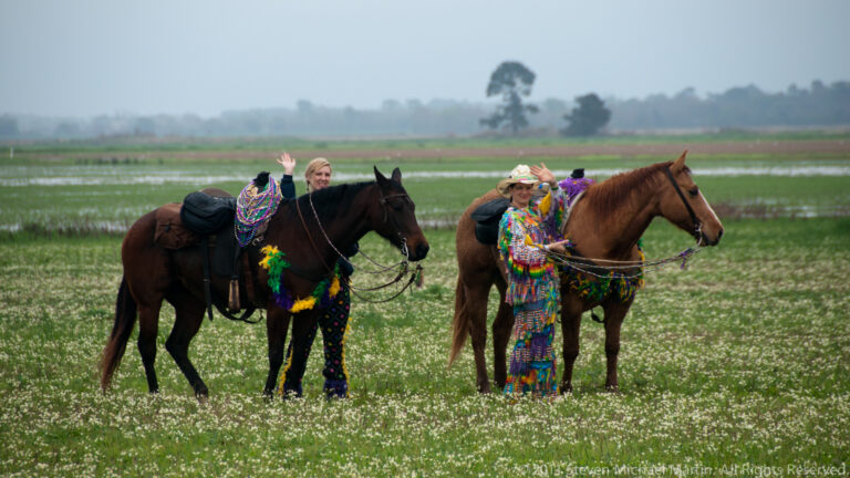 Two costumed women ride horses through a wet field, one waving toward the camera.