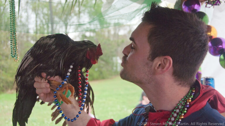 A costumed man with beads holds a black chicken close to his face beneath colorful decorations.