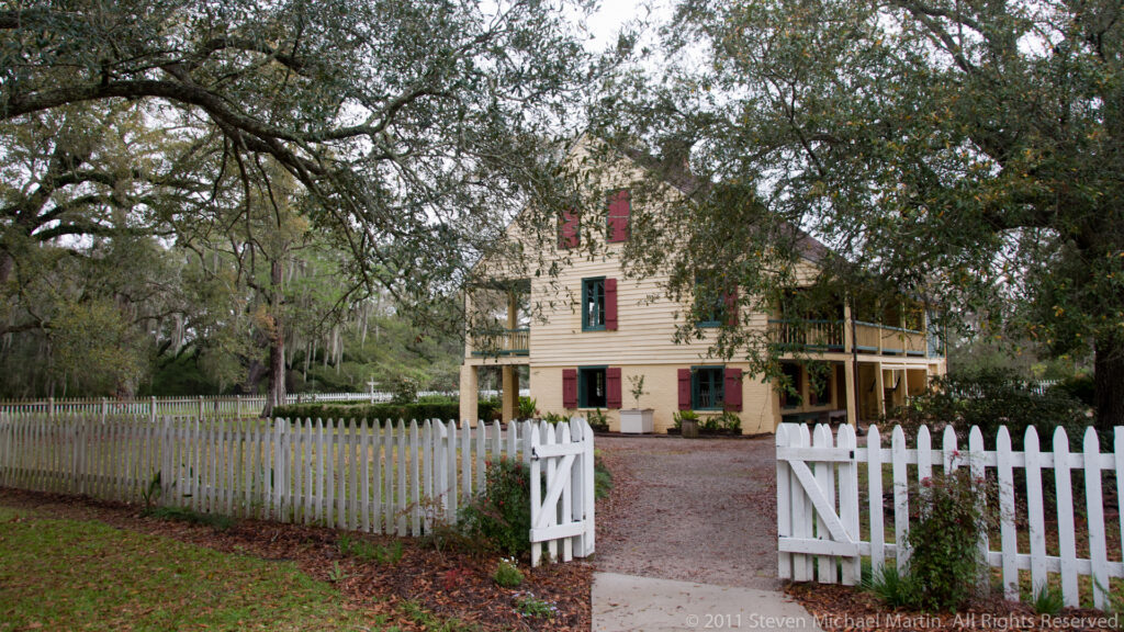 A pale two-story house with red shutters and a gallery stands behind an open white gate.