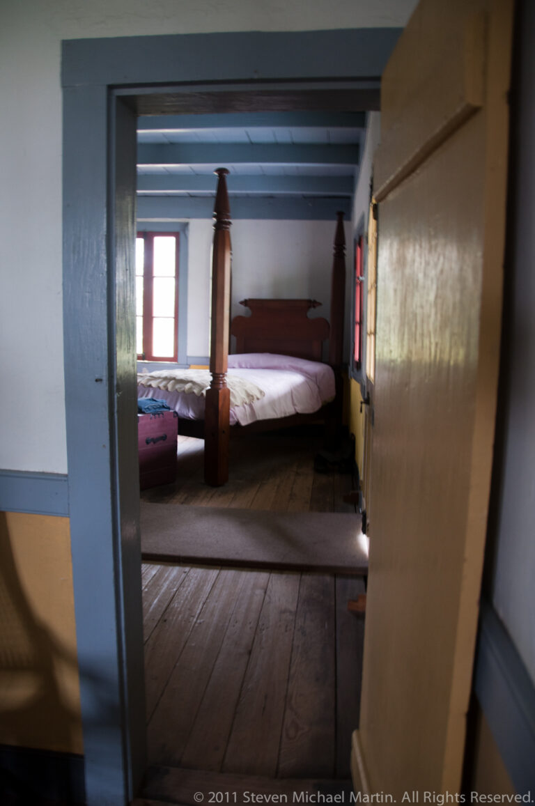 An open doorway frames a dark four-poster bed beneath blue beams and red-trimmed windows.