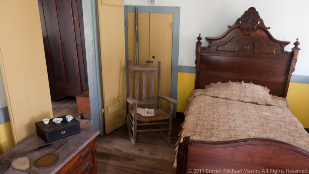 A carved wooden bed, chair, and washstand furnish a yellow room with blue-gray trim.