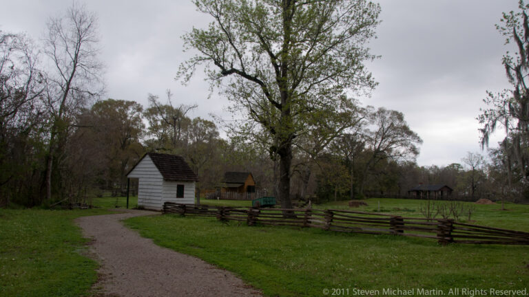 A gravel path curves past a small white outbuilding and split-rail fence on green grounds.