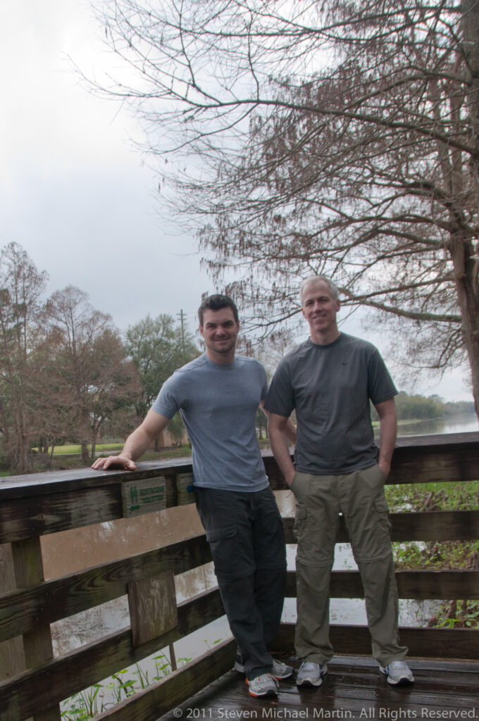 Two men stand on a wooden platform above shallow water beneath bare tree branches.