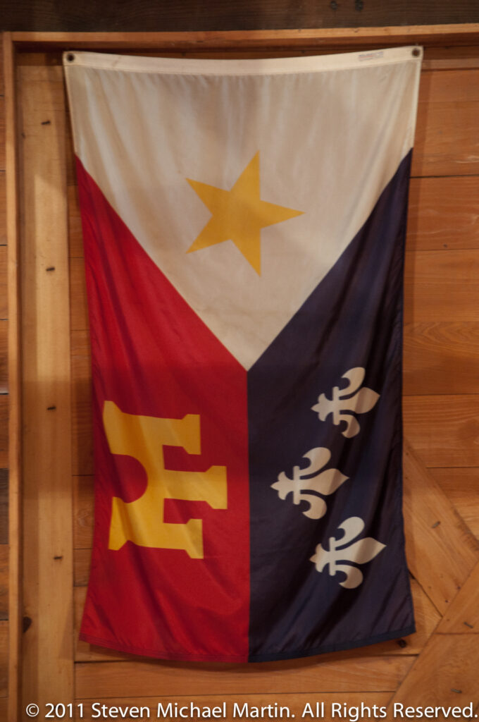 A tricolor flag with a gold star and fleur-de-lis hangs against a wooden wall.