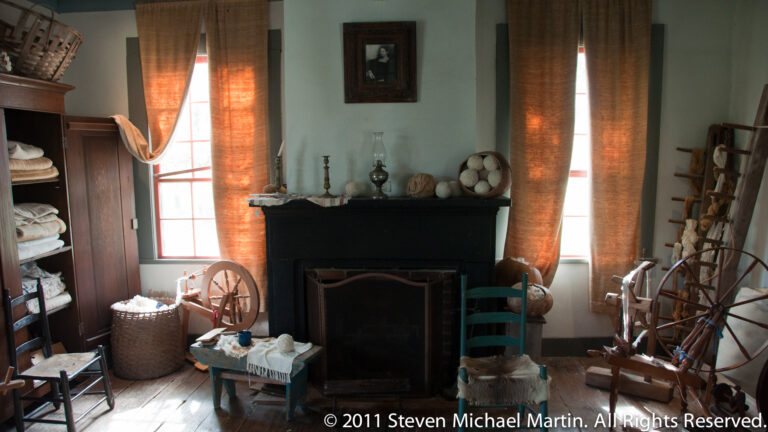 A fireplace stands between curtained windows, surrounded by a spinning wheel, baskets, chairs, and pottery.