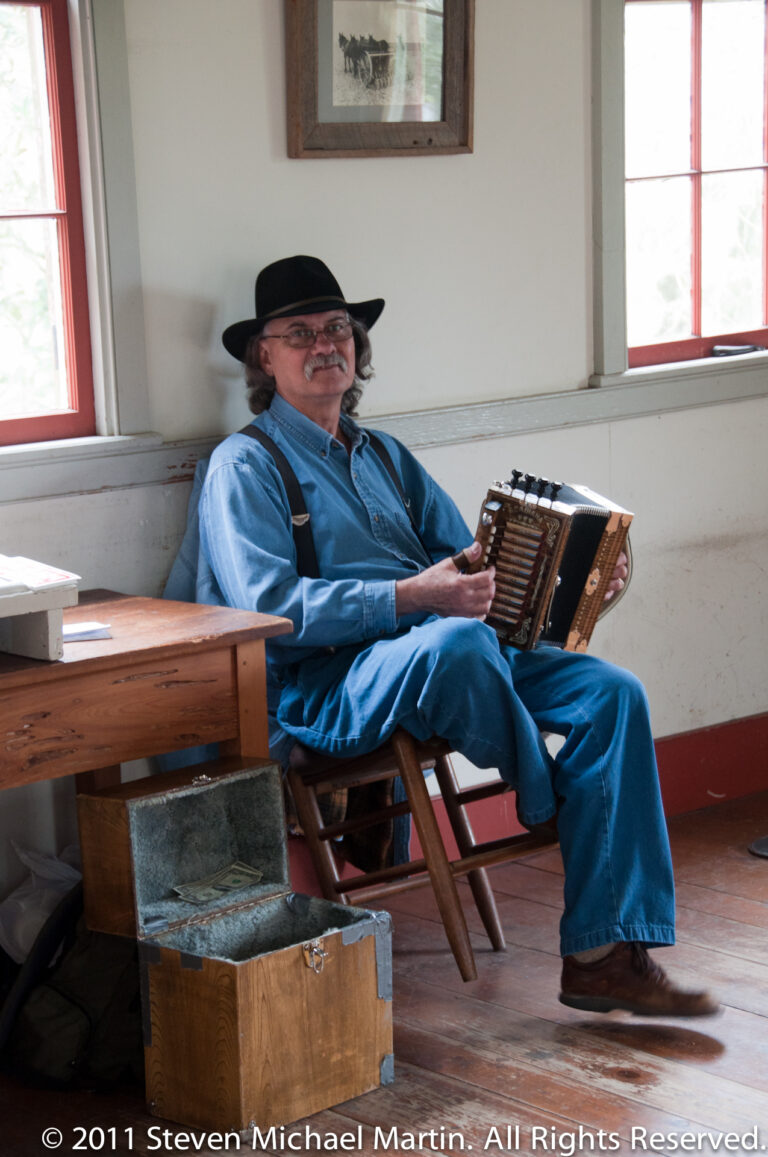 A seated man in a black hat holds an accordion beside a wooden table in a bright room.