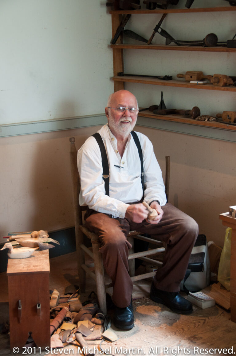A seated man in suspenders faces the camera amid shelves and floor displays of wooden tools.