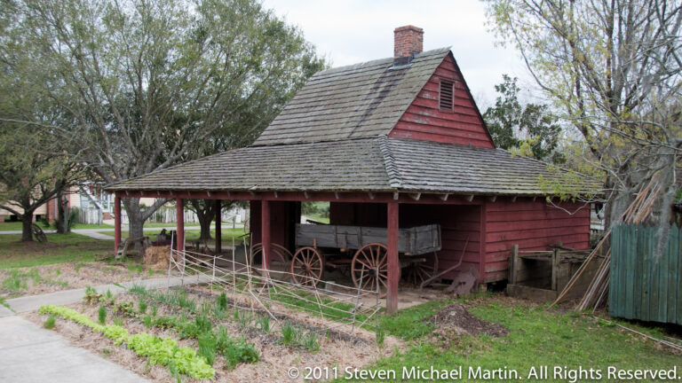 A red wooden outbuilding shelters a covered wagon beside a narrow garden bed.