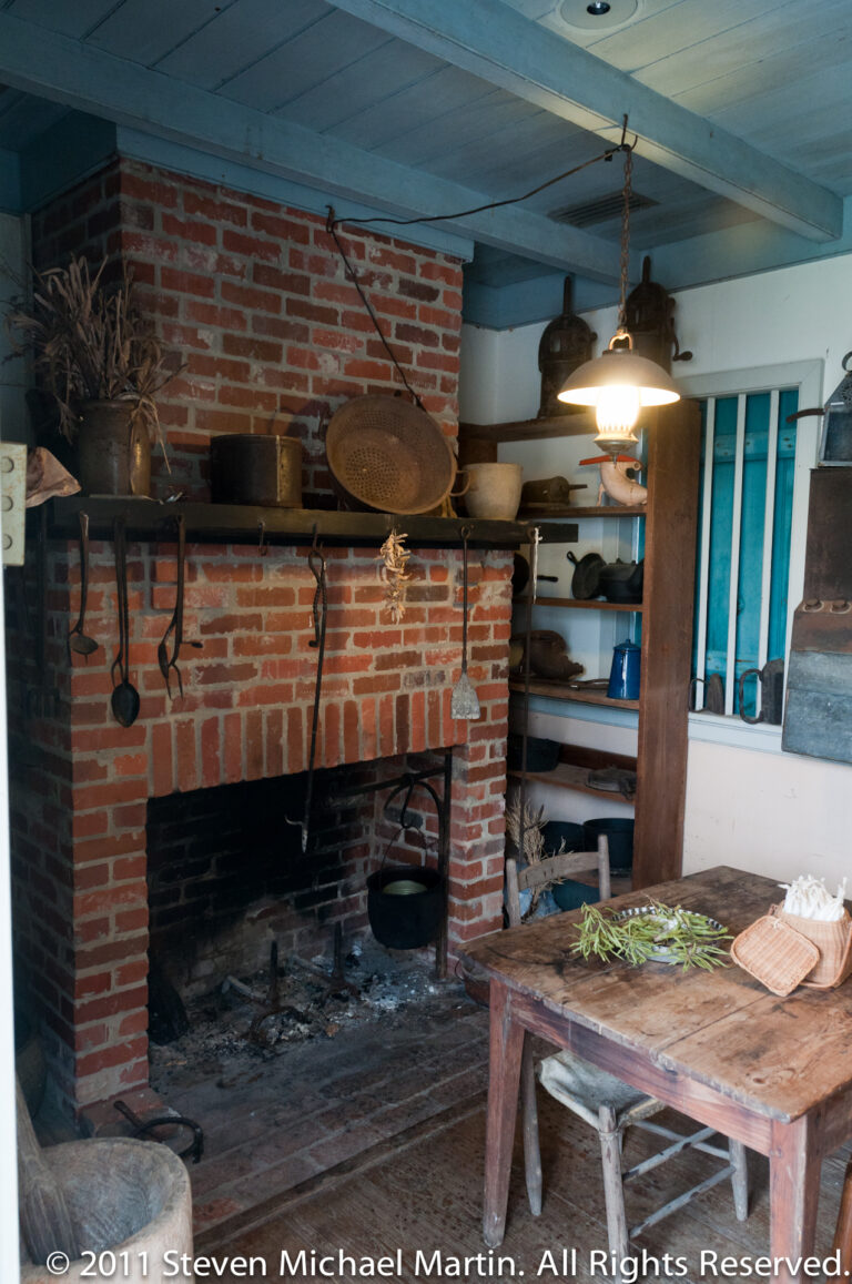 Brick hearth with iron cookware, utensils, baskets, and a worktable arranged inside a historic kitchen.