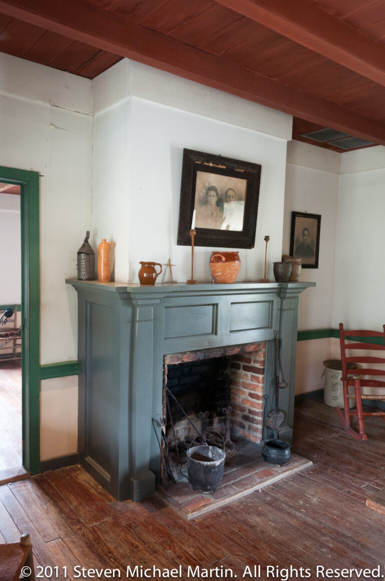 A painted mantel and brick hearth display pottery and framed pictures in a furnished room.
