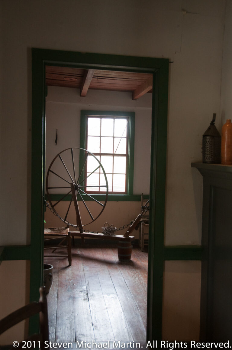 A wooden spinning wheel stands by a window, seen through a green-trimmed doorway.