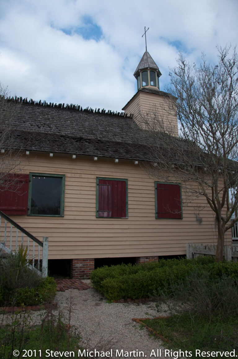 A pale raised church with red shutters and a cross-topped cupola stands beneath leafless trees.