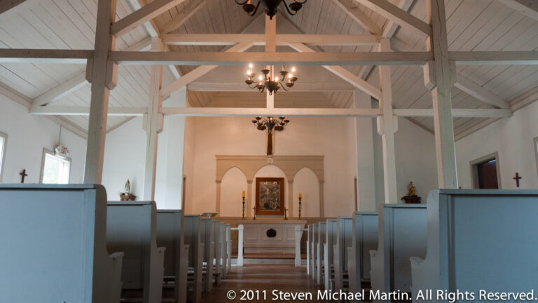 Pale blue pews flank a central aisle leading to an altar beneath exposed white roof trusses.
