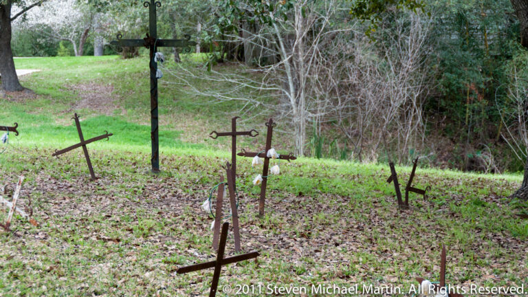 Several weathered iron crosses rise from a leaf-covered cemetery slope beneath mature trees.