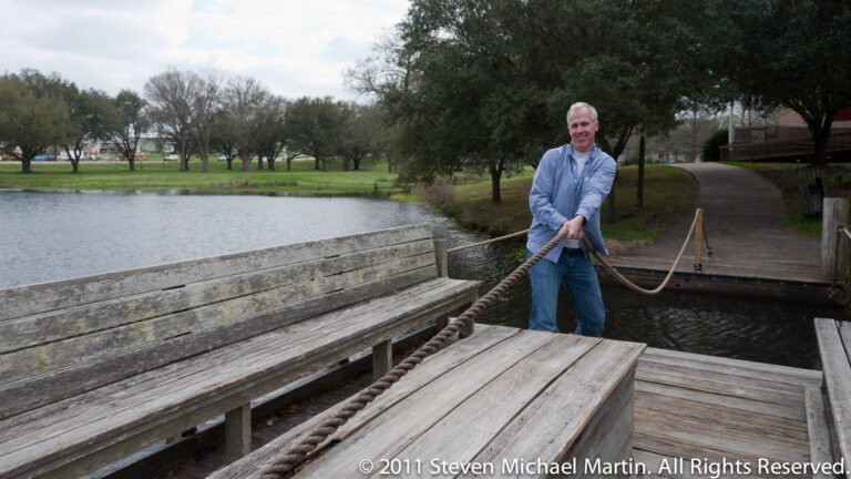 A man holds a rope on a wooden platform beside a small pond and curving park path.