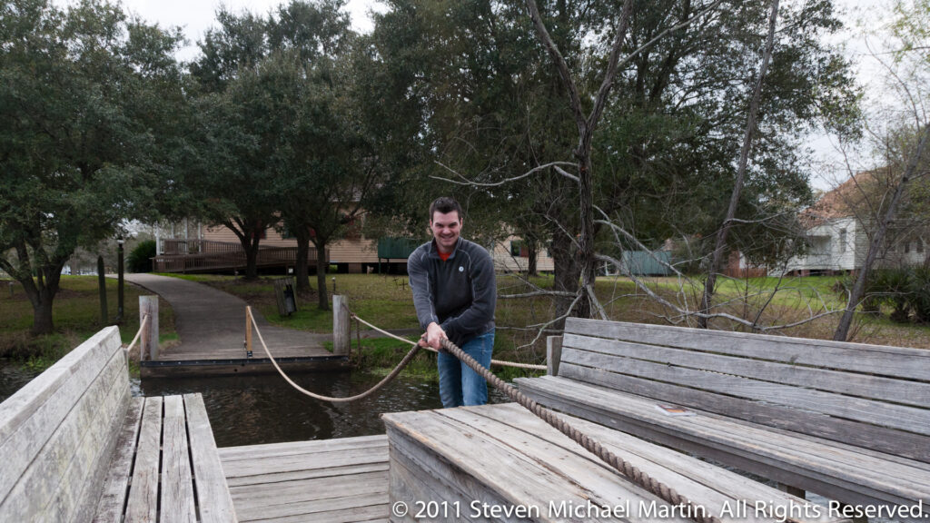 A man pulls a rope across a broad wooden dock beside a small water feature and park path.