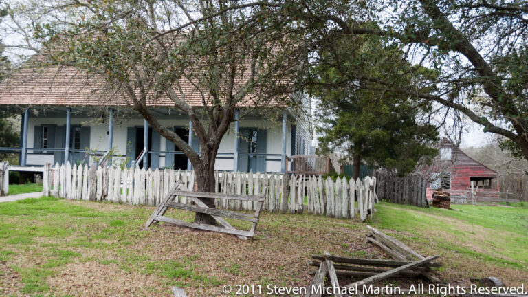 A low cottage and shaded gallery stand behind a white picket fence beneath a spreading tree.