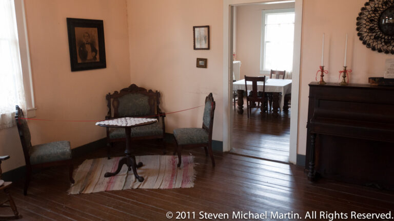 Two chairs and a small table sit in a parlor, with an open doorway leading to a dining room.