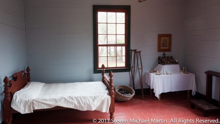 A turned-post bed sits beneath a red-trimmed window beside a display table, basket, and simple wooden furnishings.