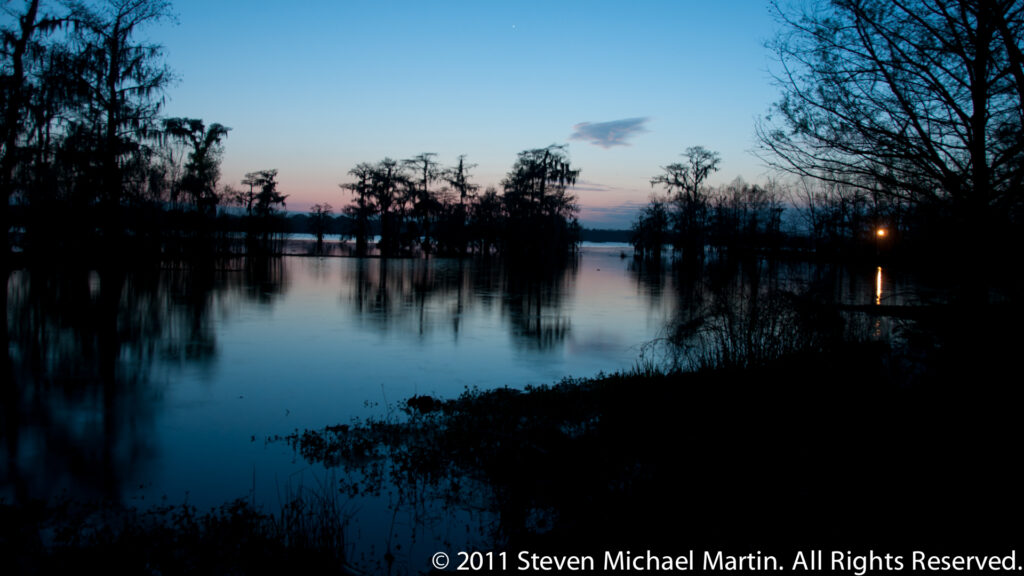 A wide blue-hour lake reflects distant cypress trees and a small amber light along the right shore.