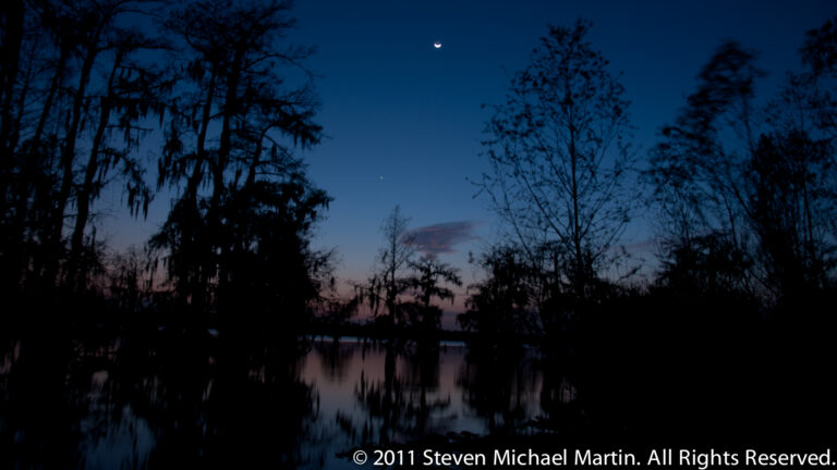 A crescent moon above silhouetted cypress trees and a narrow reflection on a Louisiana lake at twilight.