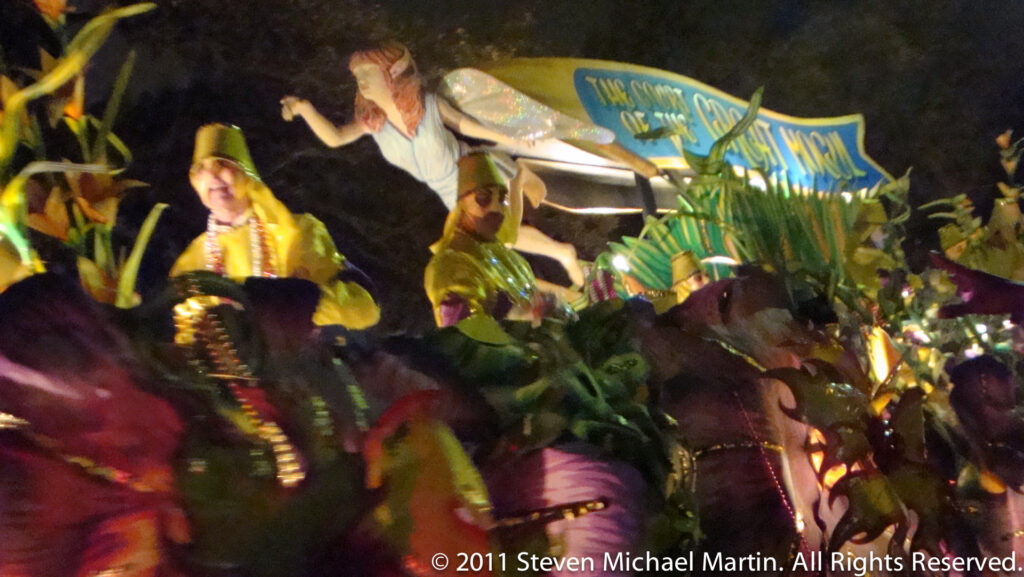 Costumed riders in yellow and green stand on a brightly illuminated float beneath a blue banner.