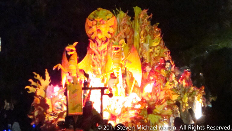 An oversized orange-red face sculpture glows above smaller figures on a nighttime parade float.