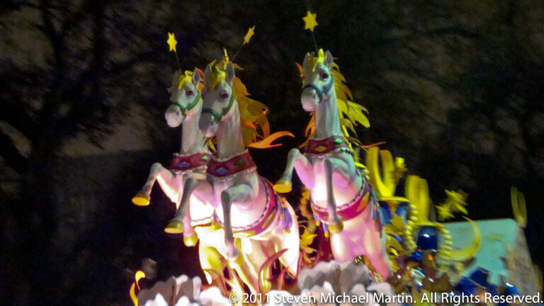 Two white sculpted horses stand on a brightly lit float with colorful feather-like decorations.